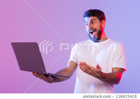 A young man with a beard and wearing a white t-shirt is holding a laptop with an excited and surprised expression on his face. The background features a gradient of purple and light blue 116615428