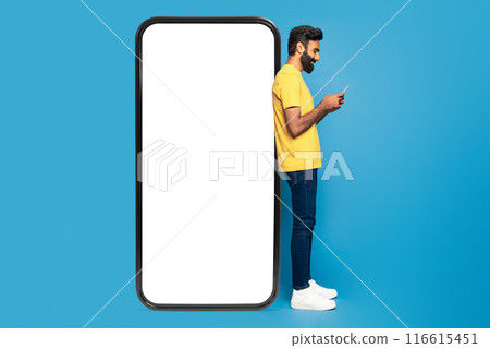 A young Indian man wearing a yellow shirt and jeans is engrossed in using his smartphone. He is standing against a giant phone frame with a blank white screen, set against a vivid blue background 116615451