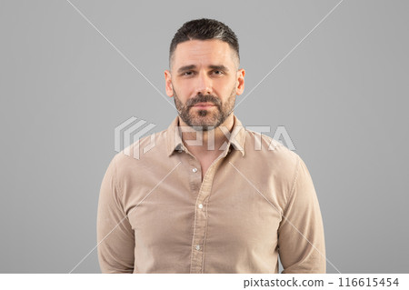 A man with a short haircut and beard is standing against a plain gray background, wearing a beige shirt and looking directly at the camera. The setting appears to be indoors with even lighting. A man with a short haircut and beard is standing against a plain gray background, wearing a beige shirt and looking directly at the camera. The setting appears to be indoors with even lighting. 116615454
