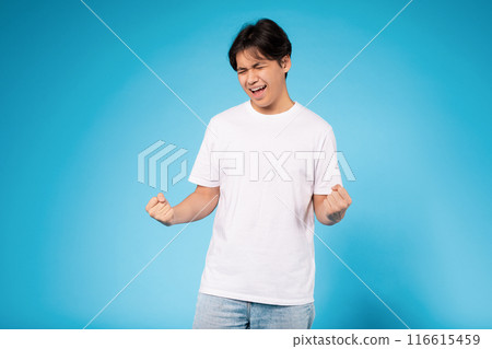 Asian young guy wearing a white t-shirt and light jeans clenches his fists in celebration against a vibrant blue backdrop. The expression on his face reflects joy and excitement. Asian young guy wearing a white t-shirt and light jeans clenches his fists in celebration against a vibrant blue backdrop. The expression on his face reflects joy and excitement. 116615459