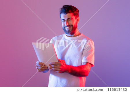 A man wearing a white t-shirt is shown holding a tablet device in his hands. He appears focused on the screen, engaged in technology. A man wearing a white t-shirt is shown holding a tablet device in his hands. He appears focused on the screen, engaged in technology. 116615480