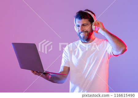 A man in a white t-shirt is standing while holding a laptop and wearing headphones. He seems to be enjoying the music in a room illuminated by neon lighting, creating a modern and vibrant atmosphere. A man in a white t-shirt is standing while holding a laptop and wearing headphones. He seems to be enjoying the music in a room illuminated by neon lighting, creating a modern and vibrant atmosphere. 116615490