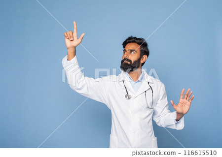 A bearded Indian doctor wearing a white lab coat and stethoscope gestures with his right hand as if pointing towards something, set against a solid blue background. 116615510