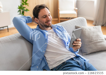 A man is casually sitting on a couch, engrossed in using a tablet device. He appears focused as he interacts with the screen, showcasing modern technology in everyday life. A man is casually sitting on a couch, engrossed in using a tablet device. He appears focused as he interacts with the screen, showcasing modern technology in everyday life. 116615535