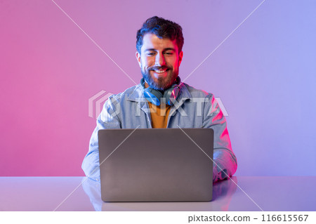 A man is sitting at a table, focused on a laptop computer in front of him. He appears to be engaged in work or study, with documents scattered around. The room is well-lit, with a neutral background. 116615567
