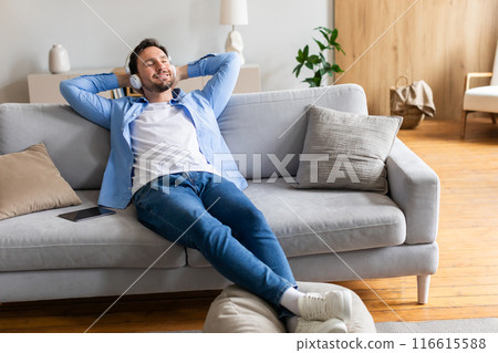 A man is comfortably seated on a couch in his living room, appearing relaxed and at ease. The room is well-lit with natural light, and the man seems to be enjoying a moment of rest and relaxation. 116615588