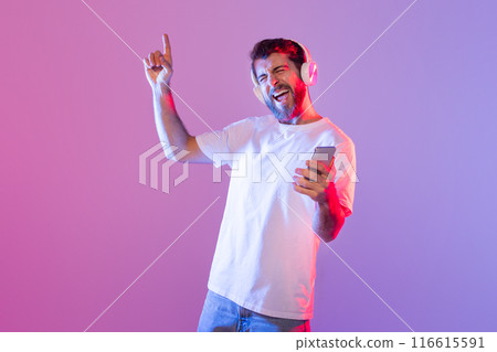 A man wearing headphones and a white t-shirt is joyfully dancing and singing while holding a smartphone. The brightly lit indoor setting features a vibrant purple and pink background 116615591