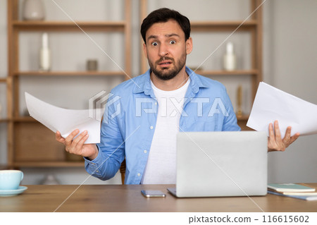 A man with a surprised expression is seated at a desk in a home office, holding papers in his hands. A laptop is open in front of him, suggesting a moment of unexpected news or work-related shock. 116615602