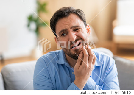 A man in discomfort is seated on a couch, holding his face in pain due to a toothache. His expression reveals his suffering as he tries to alleviate the ache. 116615610