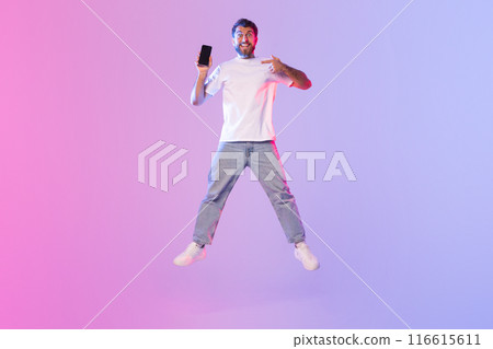 A young man dressed in a white t-shirt and jeans energetically jumps while holding and pointing at his smartphone. The backdrop features an eye-catching gradient of pink and blue 116615611