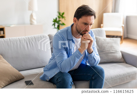 A man is seated on a couch, deep in thought, with his hand resting on his chin. He appears contemplative and engaged in introspection. A man is seated on a couch, deep in thought, with his hand resting on his chin. He appears contemplative and engaged in introspection. 116615625