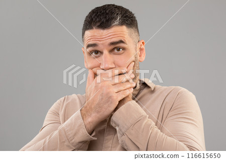 A man covers his mouth with his hands, possibly to stifle a cough, sneeze, or yawn. His hands are pressed firmly against his lips, indicating a deliberate action. 116615650
