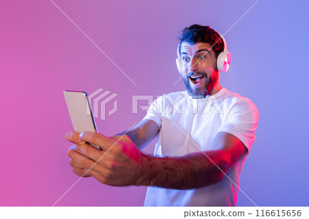 A bearded man wearing headphones and a white t-shirt is energetically using his smartphone during the night. The background is lit with vibrant blue and pink colors, highlighting his excitement. 116615656