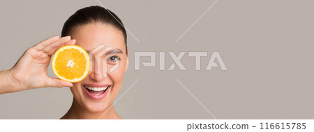 Pretty young woman holding orange half over her eye and smiling, colorful studio background, panorama with copy space 116615785
