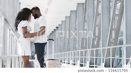 African American couple stands close together in an airport terminal, embracing before their departure. They are both dressed in white, and the man is holding a suitcase, copy space African American couple stands close together in an airport terminal, embracing before their departure. They are both dressed in white, and the man is holding a suitcase, copy space 116615795