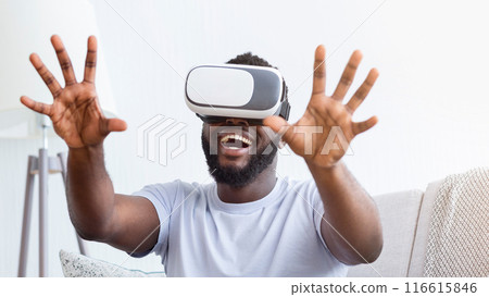 African American man wearing a VR headset is seated on a couch, both hands outstretched, as he laughs in delight at something in the virtual world. African American man wearing a VR headset is seated on a couch, both hands outstretched, as he laughs in delight at something in the virtual world. 116615846