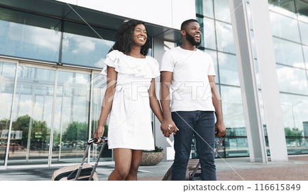 A happy African American couple walks out of an airport, each pulling a suitcase on wheels. They are both smiling and holding hands. A happy African American couple walks out of an airport, each pulling a suitcase on wheels. They are both smiling and holding hands. 116615849