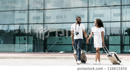 African American couple walking through an airport terminal with their luggage. They are holding hands, smiling, and appear to be in a good mood, copy space 116615850