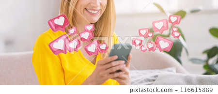 A woman sits in a living room, smiling while looking at her smartphone. Pink hearts with white centers float around her, indicating she's receiving positive feedback or likes on social media, cropped 116615889