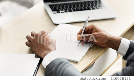 A person's hand writes in a notebook while sitting at a desk, a laptop in the background. A person's hand writes in a notebook while sitting at a desk, a laptop in the background. 116616131