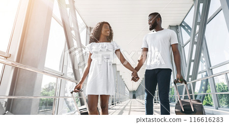 Black couple walks hand-in-hand through an airport terminal, smiling and happy. They are both pulling wheeled luggage and look excited for their upcoming vacation, copy space, sun flare 116616283