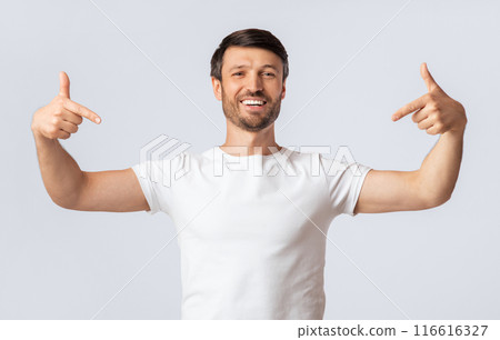 A man in a white t-shirt stands against a white background with his arms outstretched, pointing to himself with a big smile on his face. 116616327