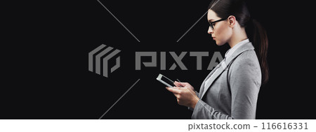 A young businesswoman in a gray suit uses her smartphone against a black backdrop. A young businesswoman in a gray suit uses her smartphone against a black backdrop. 116616331