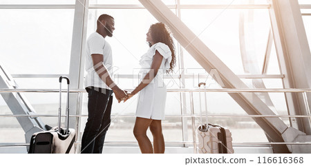 African American couple is standing in an airport terminal, holding hands and looking at each other. They are smiling, and it seems like they are about to depart on a trip together, copy space 116616368