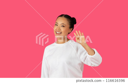Asian young woman with dark hair tied up in a bun is wearing a white sweater and standing in front of a solid pink background. She is smiling and waving hello with her right hand. Asian young woman with dark hair tied up in a bun is wearing a white sweater and standing in front of a solid pink background. She is smiling and waving hello with her right hand. 116616396