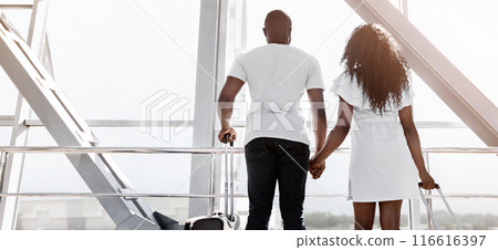 Black couple, dressed in casual clothing, stands at an airport gate, facing away from the camera and gazing towards a window. They are holding hands, copy space Black couple, dressed in casual clothing, stands at an airport gate, facing away from the camera and gazing towards a window. They are holding hands, copy space 116616397