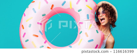 Black woman wearing a straw hat and sunglasses smiles and holds up a giant, pink inflatable float shaped like a donut. The float is covered in colorful sprinkles Black woman wearing a straw hat and sunglasses smiles and holds up a giant, pink inflatable float shaped like a donut. The float is covered in colorful sprinkles 116616399