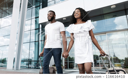 A happy Black couple, dressed in casual white attire, walks hand-in-hand towards a car, pulling their luggage behind them, outside of an airport terminal. They appear to be excited for their trip 116616410