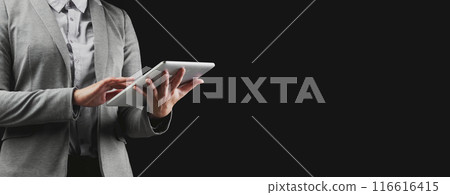 A businesswoman in a grey suit uses a tablet against a black background. 116616415