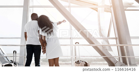 African American couple stands near an airport window, gazing out at the view, while holding hands and waiting for their flight. The woman is pointing at something out of frame, copy space 116616416
