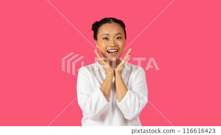 Asian young woman with dark hair in a white shirt is smiling broadly and holding her hands to her cheeks as if in surprise, standing in front of a solid pink background. Asian young woman with dark hair in a white shirt is smiling broadly and holding her hands to her cheeks as if in surprise, standing in front of a solid pink background. 116616423