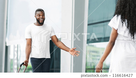 Black man with luggage is standing in a modern buildings glass-walled entrance, smiling and greeting a woman who is walking towards him. Black man with luggage is standing in a modern buildings glass-walled entrance, smiling and greeting a woman who is walking towards him. 116616427
