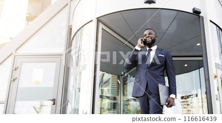 Business Workspaces For Rent. African American Businessman Talking On Cellpone Against Entrance To Modern Coworking Office Building. Low-Angle 116616439