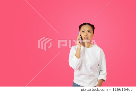 Asian woman with her hair in buns is talking on a cell phone, and she has a confused expression on her face. The background is a solid pink color, copy space Asian woman with her hair in buns is talking on a cell phone, and she has a confused expression on her face. The background is a solid pink color, copy space 116616463
