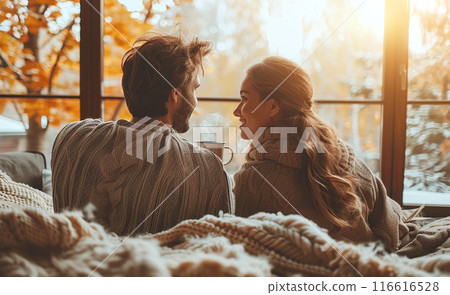 A couple sits on a couch near a window, enjoying a warm drink on a cold day. The sun is setting and they are wrapped in blankets, looking at each other and smiling. 116616528