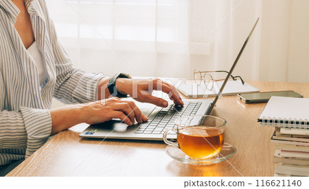 Female hands typing on a laptop with cup of tea, glasses and notepads on a wooden table Female hands typing on a laptop with cup of tea, glasses and notepads on a wooden table 116621140