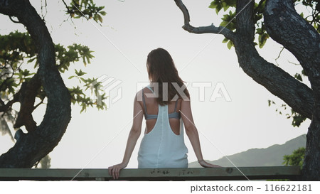 A woman wearing a white dress is seated on a wooden fence, her legs dangling in the air. She gazes into the distance, with a serene expression on her face. A woman wearing a white dress is seated on a wooden fence, her legs dangling in the air. She gazes into the distance, with a serene expression on her face. 116622181