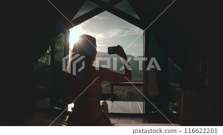 Female take photo of sunset mountain landscape from resort room villa window. Girl in bikini relax in holiday exotic wild nature hut. Outdoor lifestyle travel on summer vacation back view, indoor shot 116622201