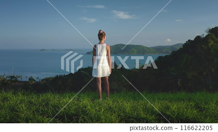 Beautiful woman in white dress stands on fresh green grass hill. Ocean wild tropical islands in background. Tranquility moment and beauty in exotic Thailand Samui paradise, summer holiday vacation 116622205