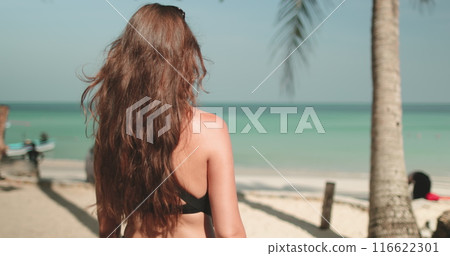 Tourist woman walking on tropical beach with palm trees, white sand beach and fishing boats. Long hair brunette girl back view. Tourism, holiday, travel to exotic Thailand island. Slow 116622301