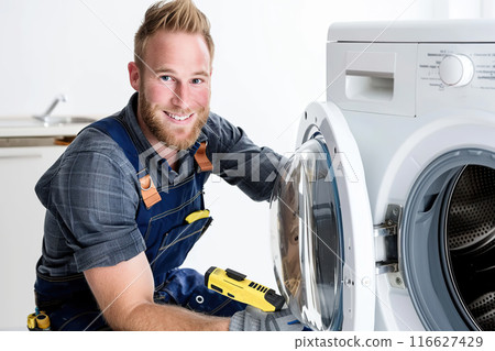Repairman and washing machine. Young handsome smiling repair man in worker suit with professional tools fixing a laundry washer in bathroom at home. Maintenance engineer plumber repairs clothes washer 116627429