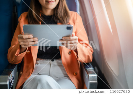 Close-up of a woman in an airplane seat using a tablet, dressed in an orange blazer and white pants, with sunlight streaming in. 116627620