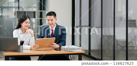 Two business professionals in a modern office setting, collaborating and discussing documents at a wooden desk. 116627625