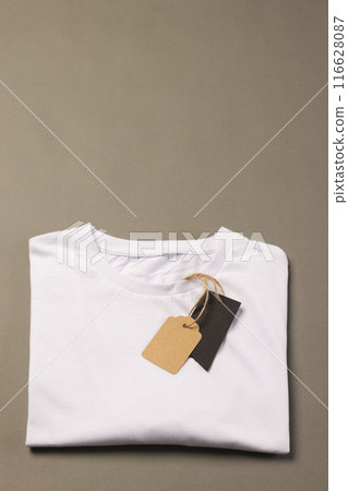 A folded white t-shirt with a blank tag attached to it lies on a neutral background, with copy space A folded white t-shirt with a blank tag attached to it lies on a neutral background, with copy space 116628087