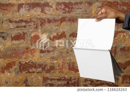 Biracial man holding blank brochures against a brick wall, with copy space 116628263