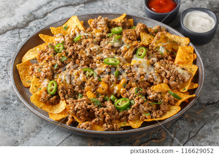 Traditional nachos with ground beef, cheddar cheese and jalapeno pepper served with sauces closeup on the plate. Horizontal Traditional nachos with ground beef, cheddar cheese and jalapeno pepper served with sauces closeup on the plate. Horizontal 116629052